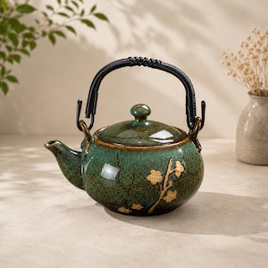 Puede incluir: Una tetera redonda de cerámica con un esmalte moteado en verde oscuro y marrón, que presenta un diseño en relieve de una rama floral de color claro. La tetera tiene una tapa a juego y un asa de alambre negro enrollado en los laterales.