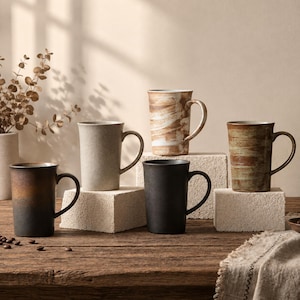 Può includere: Una collezione di cinque tazze in ceramica con varie finiture, tra cui un gradiente marrone scuro, un grigio chiaro solido, un motivo marmorizzato bianco e marrone, un nero opaco solido e un verde oliva testurizzato con pennellate orizzontali marroni.