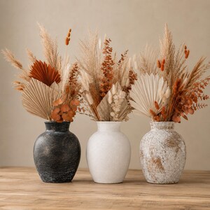 Può includere: Tre vasi arrotondati con finiture nera, bianco opaco e bianco anticato, ciascuno riempito con una composizione di fiori secchi con erba della pampa beige, eucalipto color ruggine e foglie di palma a ventaglio in tonalità ocra e arancio.