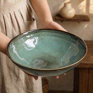 May include: A wide, round ceramic bowl featuring a reactive glaze with a gradient of deep teal and seafoam green. The interior surface has a speckled texture, while the outer rim is finished with a dark, earthy brown tone that contrasts with the lighter centre.