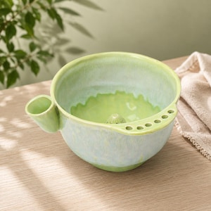 May include: A light green ceramic shaving bowl with a glossy finish. The bowl features a built-in brush holder on the side, a series of small holes along the rim for razor storage, and a textured centre point for creating a rich shaving lather.