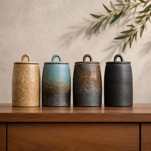 May include: Four cylindrical ceramic canisters with loop-handled lids. The first is textured tan, the second features a blue-to-brown gradient, the third has a metallic bronze-to-charcoal finish, and the fourth is a solid matte black.
