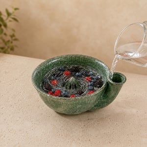 Könnte beinhalten: Eine gesprenkelte grüne Keramikschale mit einer zentralen Zitruspresse und einem seitlichen Ausguss, gefüllt mit Wasser, Blaubeeren und Himbeeren. Ein transparenter Messbecher gießt Wasser in den Ausguss der Schale und erzeugt einen Wirbel.