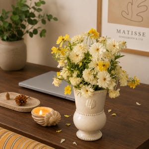 May include: A cream-coloured ceramic vase with a beaded rim and a small embossed face detail, filled with a bouquet of yellow and white cosmos flowers. The vase sits on a dark wooden surface alongside a shell-shaped candle holder with a lit flame.