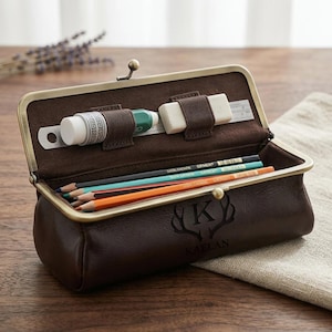 May include: A dark brown leather pencil case with a brass-toned metal frame and clasp. The front is embossed with a letter K and antlers above the name KAELAN. The interior features brown fabric lining, elastic loops holding a white eraser, a green marker, and a metal ruler, with three colored pencils resting inside.