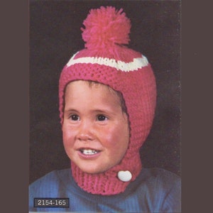 以下が含まれることがあります： ピンクのニット帽に白いストライプとピンクのポンポンが付いています。帽子には耳と首を覆うフラップが付いています。帽子は青いシャツを着た子供にかぶせられています。画像には「2154-165」というテキストが含まれています。