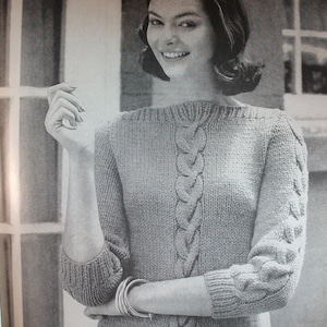 May include: A close-up of a woman wearing a gray knitted sweater with a cable knit design. The sweater has a round neckline and long sleeves. The woman is wearing several bracelets on her wrist.