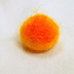May include: A small, round, handmade felt ball in shades of orange and yellow. The felt ball has a fuzzy texture and is set against a white background. This item could be used for crafting or as a decorative element.