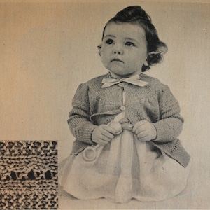 Puede incluir: Una foto en blanco y negro de un niño pequeño que lleva un cárdigan y un vestido de punto. El niño sostiene una cuchara de madera en sus manos.