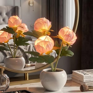 May include: A decorative lamp shaped like a cluster of light-up pink peony flowers with green leaves, potted in a smooth white ceramic bowl. The flowers emit a soft, warm glow, and the entire arrangement is reflected in a circular gold-framed mirror.