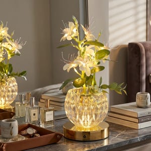 May include: A decorative table lamp featuring a clear, textured glass vase base with a gold-toned circular stand. The lamp is illuminated with warm LED lights integrated into a cluster of white artificial flowers and green leaves.