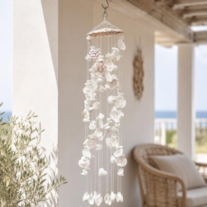 Peut inclure: Un carillon éolien mobile suspendu composé de nombreux coquillages blancs et blanc cassé fixés par de fines ficelles blanches à un cadre circulaire. Les coquillages varient en taille et en forme, créant un agencement vertical en cascade.