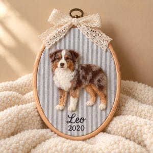 May include: An oval wooden embroidery hoop containing a needle-felted Australian Shepherd dog on grey and white striped fabric. A cream-coloured lace bow is attached to the top. The name Leo and the year 2020 are embroidered in black thread below the dog.