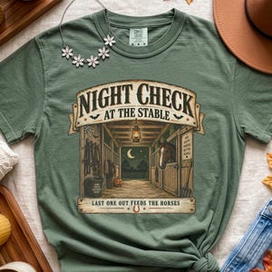 May include: A sage green short-sleeve t-shirt featuring a graphic of a stable interior with a horse, a lantern, and the text NIGHT CHECK AT THE STABLE LAST ONE OUT FEEDS THE HORSES. The shirt has a crew neckline and a tag labeled Comfort Colors.