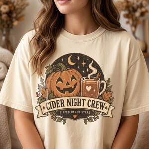 May include: A cream-colored t-shirt featuring a graphic of a jack-o'-lantern, a steaming mug with a heart, and autumn leaves under a crescent moon. The text reads CIDER NIGHT CREW and SIPPED UNDER STARS, framed by a decorative banner design.