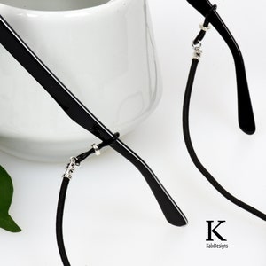 May include: Black eyeglasses with a black cord and silver accents. The cord is attached to the frames with small silver connectors. The eyeglasses are set against a white background. The text "K KaixDesigns" is in the lower right corner.