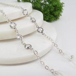 May include: A pair of clear crystal eyeglass chains with silver metal chain links and clear silicone ends. The chains are laying on a white surface.