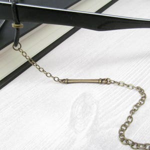 May include: A bronze chain with a long, thin bar connector. The chain is attached to a pair of black eyeglasses.
