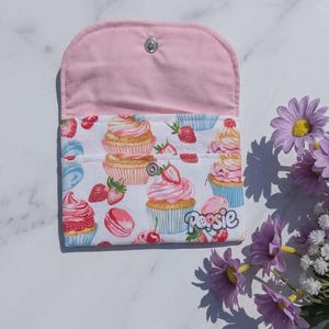 Può includere: Una pochette in tessuto con interno rosa chiaro ed esterno bianco caratterizzato da un motivo ripetuto di cupcake, fragole e macaron. La parola "Popsie" è stampata in corsivo sul davanti. Include una chiusura a bottone a pressione.