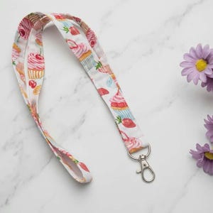 Può includere: Un cordino in tessuto con un motivo ripetuto di cupcake, fragole e ciliegie nei toni del rosa, rosso e azzurro. Il cordino è dotato di un moschettone girevole in metallo color argento fissato all'estremità per agganciare vari oggetti.