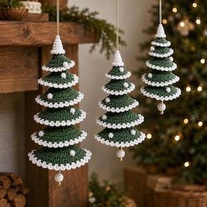 May include: Three hanging Christmas tree ornaments crafted from dark green yarn with white crocheted edges and small white pom-poms. Each tree features a pointed top, tiered layers, and a single white bead dangling from the bottom on a gold string.