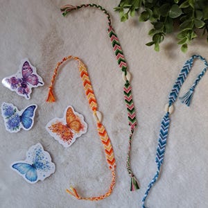 Könnte beinhalten: Drei gewebte Freundschaftsarmbänder mit Chevron-Mustern in Orange, Grün und Blau, jeweils akzentuiert mit einer kleinen weißen Muschelperle. Neben den Armbändern befinden sich vier schmetterlingsförmige Aufkleber mit Blumen- und Aquarellmotiven.