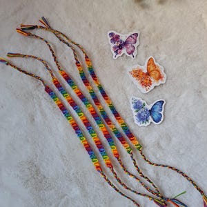 Könnte beinhalten: Vier handgefertigte Freundschaftsarmbänder mit einem leuchtenden, regenbogenfarbenen Knüpfmuster und geflochtenen Enden, begleitet von drei einzelnen Schmetterlingsaufklebern, einer mit violetten Blumenflügeln, einer orange und einer blau mit Blumenakzenten.