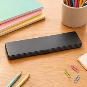 Könnte beinhalten: Ein rechteckiges, mattschwarzes Stifteetui aus Metall mit abgerundeten Ecken und einem Klappdeckel. Das Etui liegt horizontal auf einer Holzoberfläche, begleitet von bunten Notizbüchern, einem Stiftehalter und mehreren Büroklammern.