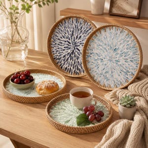May include: A collection of four round woven rattan trays featuring decorative interior patterns. Two trays are upright, displaying blue and white petal designs, while two lie flat, holding a bowl of cherries, a bread roll, and a cup of tea.