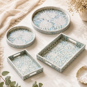 Puede incluir: Un conjunto de cuatro bandejas decorativas que incluye dos bandejas redondas y dos rectangulares con asas integradas. Cada bandeja muestra un patrón de mosaico con piezas iridiscentes en tonos azul, blanco y turquesa con motivos florales.
