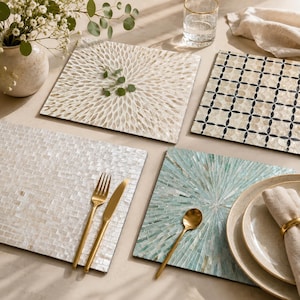 May include: Four square placemats featuring distinct designs: one with a white radial petal pattern, one with a black and white geometric grid, one with a white tiled mosaic, and one with a light blue and white sunburst mosaic pattern.