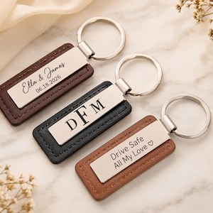 May include: Three rectangular keychains with silver-toned metal plates attached to leather-like tabs in dark brown, black, and light brown. The plates feature engraved text: Ella & James 06.18.2026, DFM, and Drive Safe All My Love with a heart symbol.
