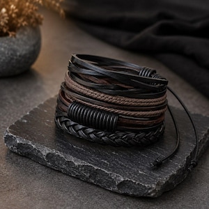 Può includere: Un bracciale multistrato caratterizzato da una combinazione di cordini in pelle nera e marrone, una treccia in pelle nera e un componente centrale a forma di perla cilindrica nera. Il design include sottili lacci in pelle che si estendono.