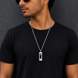 Può includere: Una collana con ciondolo rettangolare color argento caratterizzata da un design con lettera G ritagliata, sospesa a una catenina argentata abbinata. Il ciondolo ha una finitura metallica lucida e poggia su una t-shirt nera.