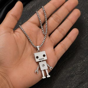 May include: A silver-toned robot pendant necklace featuring a boxy head with two circular eyes and a rectangular mouth, a square torso with a central circular detail, and articulated arms and legs, suspended from a metallic box chain.