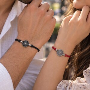 May include: Two adjustable cord bracelets featuring circular metal charms. One bracelet has a black cord with a black and silver sunburst charm, while the other has a red cord with a silver sunburst charm. Both include small silver beads.