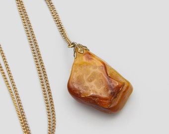 May include: A gold-colored chain necklace with a large, irregularly shaped pendant. The pendant is a mix of orange, brown, and cream colors, with a textured surface. The necklace is displayed on a white background.