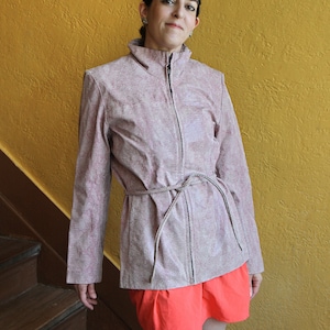 May include: A pink, textured jacket with a high collar and a front zipper. The jacket has a matching belt tied around the waist. The person is wearing a pair of orange shorts. The background is a yellow wall and a wooden staircase.