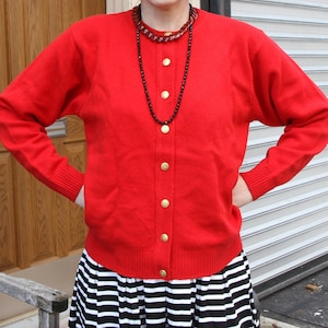 May include: A vibrant red cardigan with gold buttons, long sleeves, and a ribbed hem. It is worn with a black and white striped skirt. A black beaded necklace is also visible.