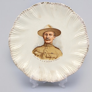 May include: A decorative, off-white plate with a scalloped edge and gold trim. The plate features a portrait of a man in a tan uniform and hat. The text "Lt. Col. R.S. Baden Powell" is printed below the portrait.