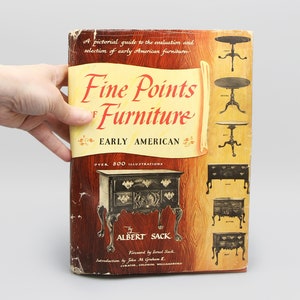 Pode incluir: Um livro vintage intitulado "Fine Points of Furniture" com o subtítulo "Early American". A capa apresenta ilustrações de móveis e o nome do autor, Albert Sack. O livro é segurado por uma mão.