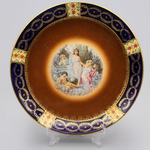 May include: Decorative ceramic plate with a brown center and a detailed border. The border is dark blue with gold accents and a repeating pattern. The center features a classical scene with figures in flowing robes.