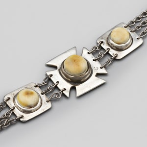 Pode incluir: Uma pulseira de prata com três elos quadrados e em forma de cruz, cada um com uma pedra redonda de cor creme. Os elos são conectados por três correntes paralelas. A joia está sobre um fundo cinza.
