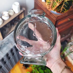 Peut inclure: Vase transparent en forme d'horloge. Le vase a un corps rond avec un motif de cadran d'horloge, comprenant des chiffres et des aiguilles. Le vase a une base courte et un large orifice. Le vase est tenu par une personne.
