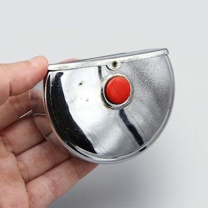 May include: A vintage, chrome-plated bicycle headlight with a bright red button. The headlight has a curved, half-moon shape and is held in a hand against a white background. The chrome finish reflects light.