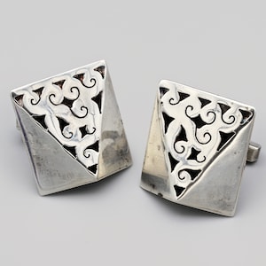 Può includere: Un paio di gemelli d'argento. Ogni gemello ha una forma quadrata con un taglio diagonale e un motivo traforato decorativo con accenti neri. I gemelli sono esposti su uno sfondo grigio chiaro. Un accessorio classico.