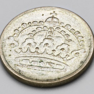 Puede incluir: Una moneda plateada con un diseño detallado de corona. La corona está intrincadamente representada con una cruz en la parte superior. La moneda tiene una apariencia ligeramente desgastada.