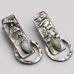 May include: A pair of silver earrings. Each earring has a textured rectangular top and a circular bottom. They feature a light green oval stone. The earrings have a hammered metal finish, giving them a rustic appearance.