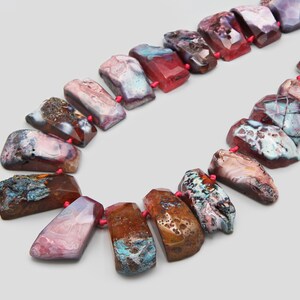 Puede incluir: Un collar con cuentas de piedra pulida de forma irregular en tonos rojos, marrones, azules y morados. Las cuentas están ensartadas con hilo rojo, creando un accesorio único y colorido.