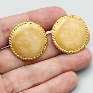 May include: A pair of round, gold-toned clip-on earrings. Each earring features a cream-colored, marbled center surrounded by a twisted rope-like border. The earrings are displayed on a light gray background.